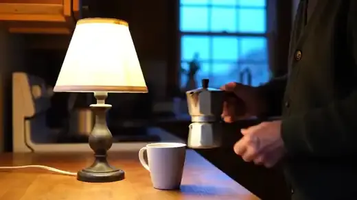 Lamplit kitchen before sunrise, a man in a green cardigan pouring coffee into a white mug